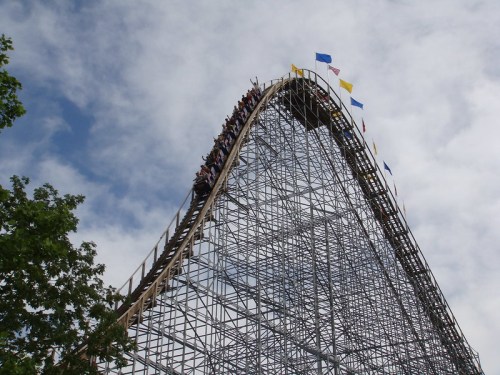 A roller coaster reaches its peak, with riders at the top and colorful flags waving against a cloudy sky.