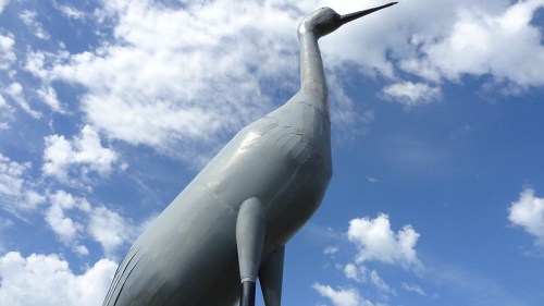 A large metallic crane sculpture stands against a blue sky with fluffy white clouds.