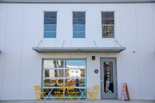 A modern coffee shop with large windows, a colorful sign, and yellow chairs outside on a sunny day.
