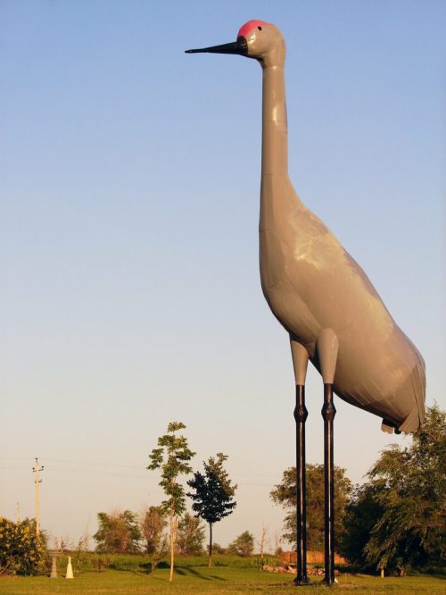 A large, gray crane statue stands tall against a clear blue sky, surrounded by trees and open land.