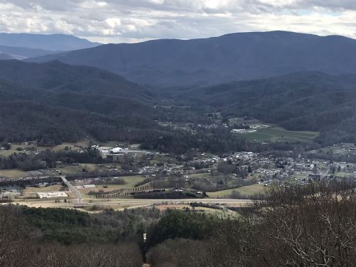 A panoramic view of a valley surrounded by mountains, with a small town and fields visible below under a cloudy sky.