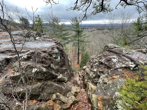 A rocky outcrop overlooks a forested valley under a cloudy sky, with trees and distant hills visible.