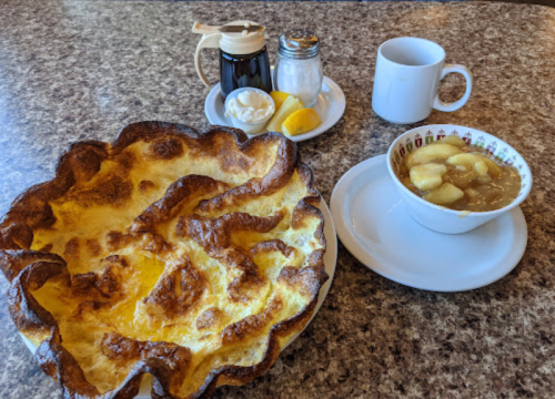 A large, golden-brown Dutch baby pancake with syrup, sugar, and a cup of apple cinnamon sauce on a table.