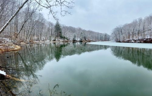 A serene lake surrounded by snow-covered trees, reflecting the cloudy sky and winter landscape.