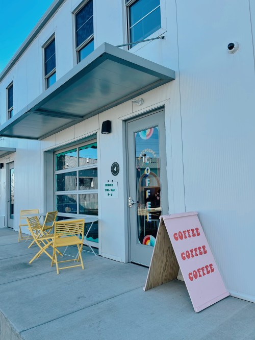 A modern coffee shop entrance with a colorful sign, yellow chairs, and a pink sandwich board displaying "COFFEE."