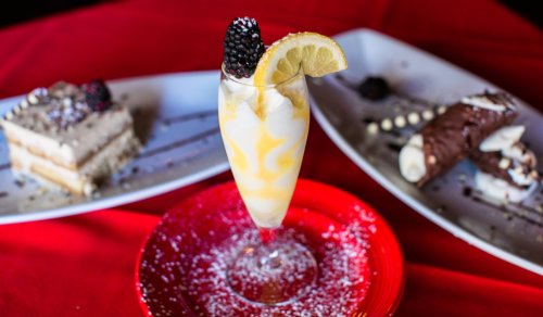 A glass of dessert with lemon and blackberry, surrounded by slices of cake and cannoli on white plates.