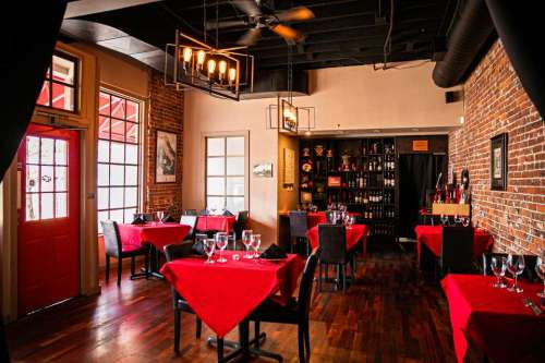 Cozy restaurant interior with red tablecloths, wooden floors, and exposed brick walls, featuring elegant lighting.