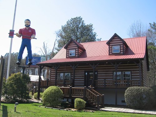 A log cabin-style building with a red metal roof and a large figure of a man on a pole nearby.
