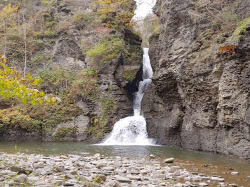 A serene waterfall cascades down rocky cliffs into a calm pool, surrounded by autumn foliage and smooth stones.