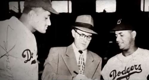 Two baseball players in Dodgers uniforms watch as a man in a suit writes notes, likely discussing a contract or agreement.
