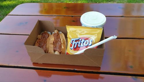 A cardboard tray with two hot dogs topped with sauce, a bag of Fritos, and a cup of drink on a wooden table.
