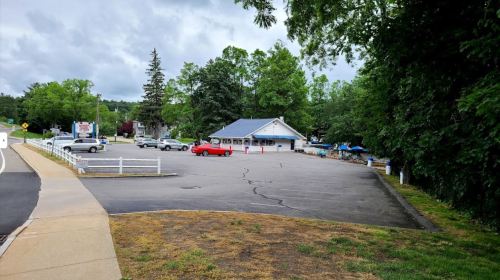 A parking lot with several cars, a building in the background, and trees lining the area under a cloudy sky.