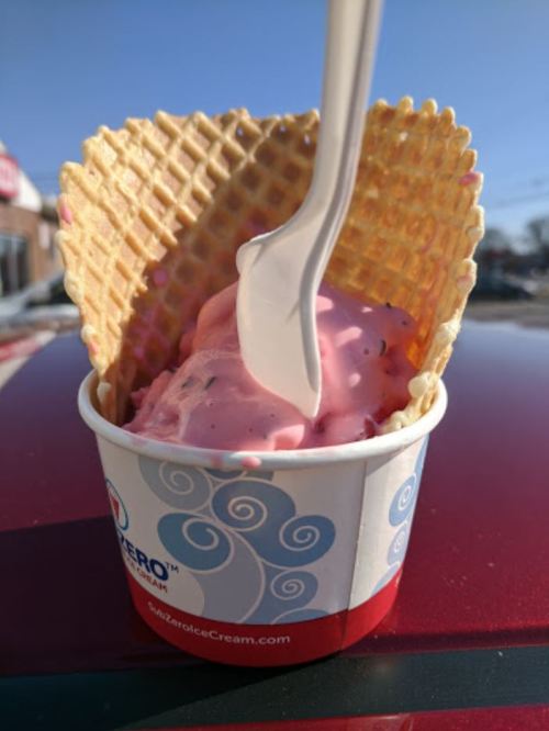 A cup of pink ice cream in a waffle cone, with a plastic spoon, resting on a red surface.