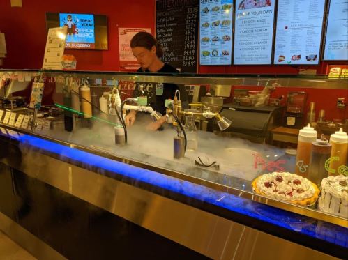A worker prepares desserts at a counter with smoke effects, surrounded by menu items and toppings.