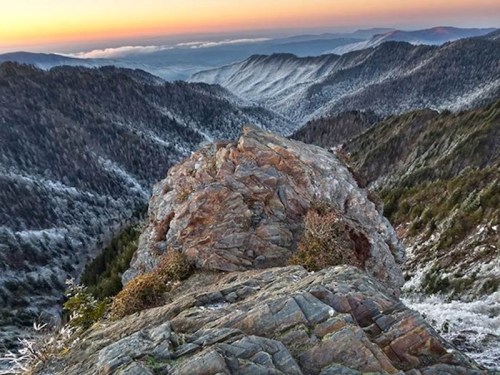 A rocky outcrop overlooks a misty mountain range at sunset, with snow-dusted peaks and a colorful sky.
