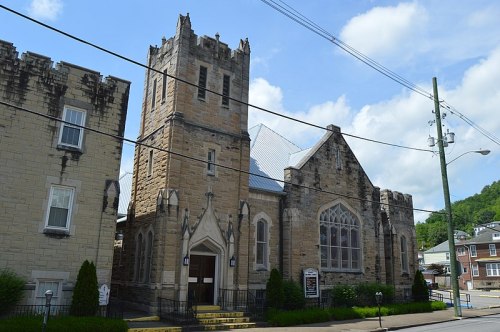 Historic stone church with a tall tower, surrounded by greenery and buildings, under a blue sky with clouds.