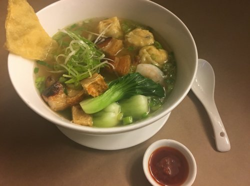 A bowl of noodle soup with greens, shrimp, and crispy toppings, served with a side of chili sauce.