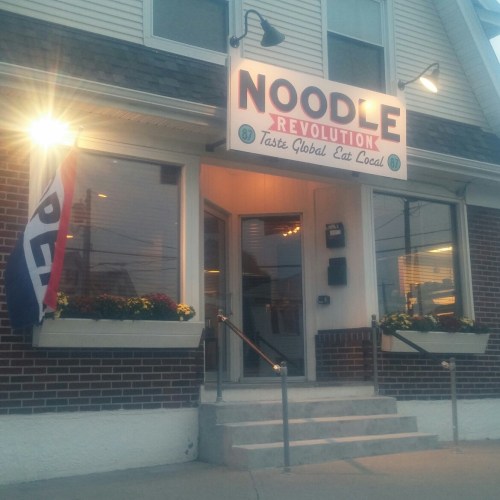 Exterior of Noodle Revolution restaurant, featuring a sign and open flag, with flower planters by the entrance.