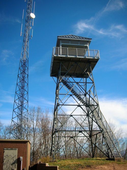 A tall metal fire tower with a staircase, next to a communication tower, against a blue sky and bare trees.