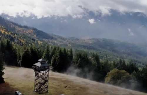 A wooden observation tower stands in a misty field, surrounded by dense forests and mountains under a cloudy sky.