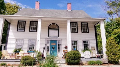 A charming white house with a blue door, large porch, and greenery, featuring a welcoming atmosphere.