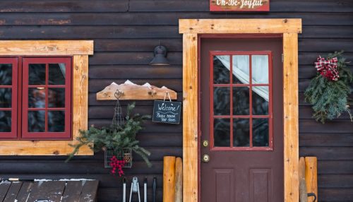 A cozy cabin entrance with a wooden door, festive decorations, and a welcome sign. Snow is visible on the roof.