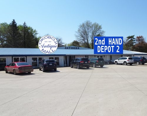 Exterior view of a mall featuring "Okoboji Mall" and "2nd Hand Depot 2" signs, with parked cars in front.
