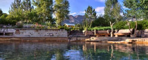 A serene outdoor hot spring surrounded by trees and mountains, with wooden seating and steps leading to the water.