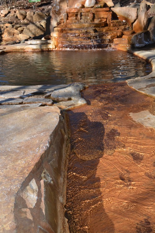 A serene hot spring with a stone pathway leading to a small waterfall and a calm pool of water.