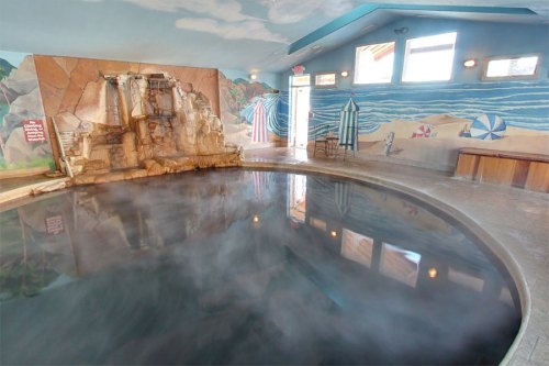 Indoor hot spring pool with a stone waterfall, surrounded by beach-themed murals and mist rising from the water.