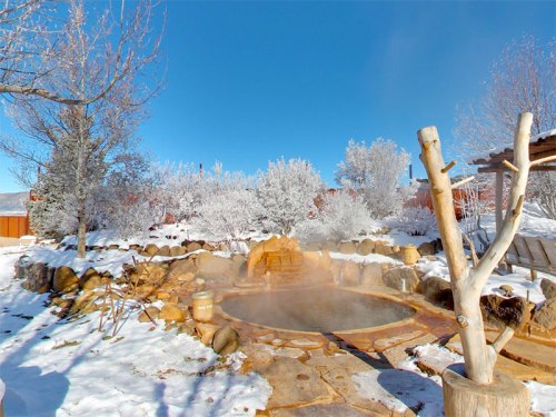 A steaming hot spring surrounded by snow-covered trees and rocks under a clear blue sky.