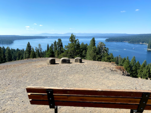 A wooden bench overlooks a serene lake surrounded by trees and mountains under a clear blue sky.