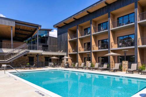 Modern hotel exterior featuring a swimming pool, lounge chairs, and a multi-level building under a clear blue sky.