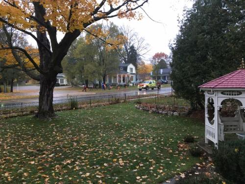 A rainy street scene with autumn leaves, a gazebo, and people walking near parked emergency vehicles.