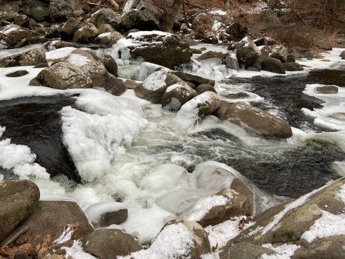 A frozen river flows over rocks, surrounded by snow and ice in a serene, winter landscape.