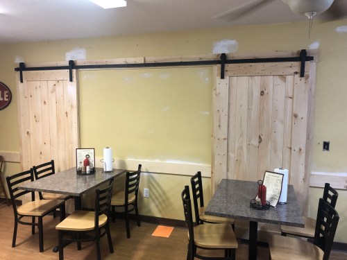 Interior of a restaurant featuring two wooden sliding doors and tables with chairs, set against a yellow wall.