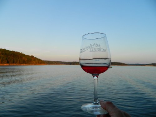 A hand holds a glass of red wine against a serene lake view at sunset.