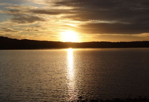 A serene sunset over a calm lake, with golden reflections on the water and dark silhouettes of distant hills.