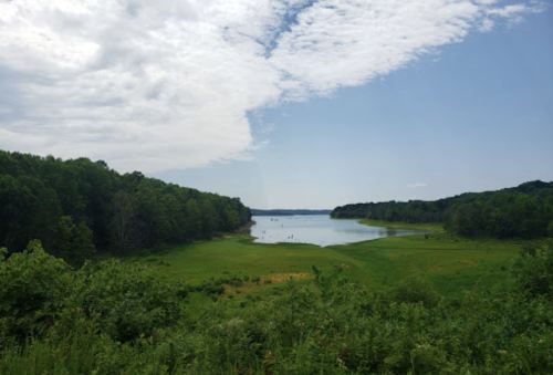 A serene lake surrounded by lush greenery under a partly cloudy sky, with a few people in the distance enjoying the water.