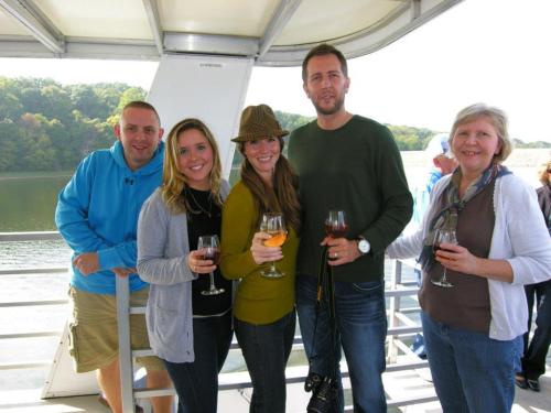 A group of five people on a boat, smiling and holding drinks, with a scenic view of water and trees in the background.