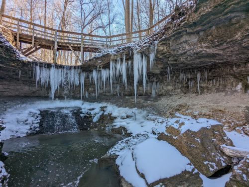 A winter scene featuring icicles hanging from rocky cliffs above a small waterfall, surrounded by snow-covered ground and trees.