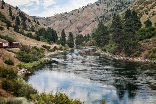 A serene river flows through a mountainous landscape, with trees lining the banks and a train passing nearby.