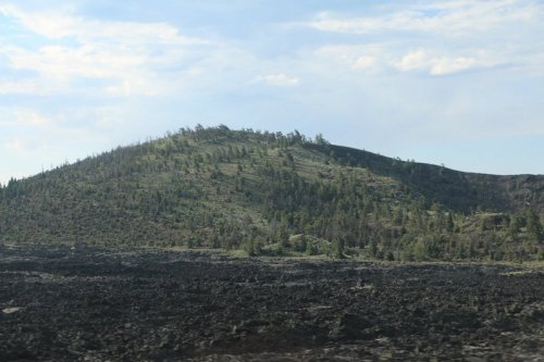 A volcanic landscape featuring a grassy hill surrounded by dark, rocky lava fields under a blue sky.