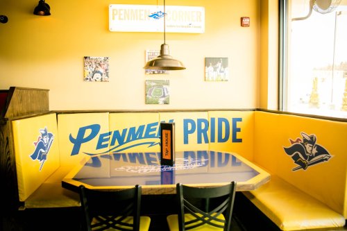 A cozy restaurant booth with yellow seating, featuring "Penmen Pride" and a table reflecting the surroundings.