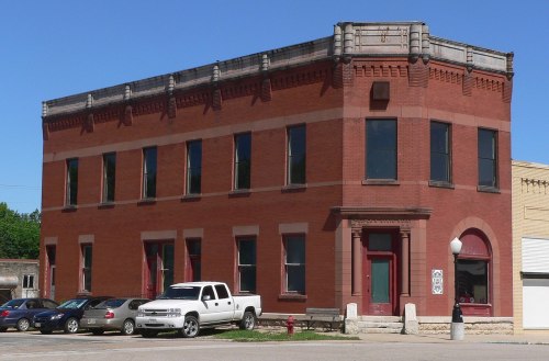 Historic red brick building with large windows, featuring a decorative cornice, and parked cars in front.
