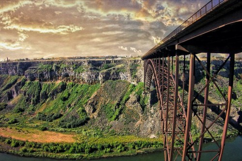 A scenic view of a bridge spanning a canyon, with lush greenery and a river below under a dramatic sky.