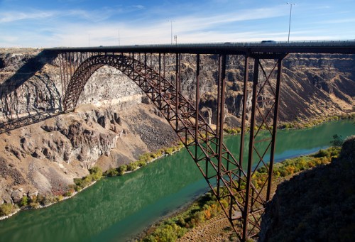 A large metal arch bridge spans a deep canyon with a green river below, surrounded by rocky terrain and sparse vegetation.