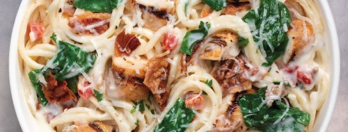 Creamy pasta with grilled chicken, spinach, tomatoes, and bacon, topped with grated cheese in a white bowl.