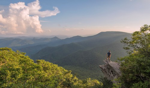 A person stands on a rocky ledge, overlooking a vast mountain landscape under a blue sky with clouds.