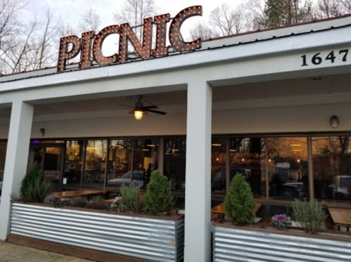Exterior of a restaurant named "PICNIC," featuring a sign with lights and large windows, surrounded by greenery.